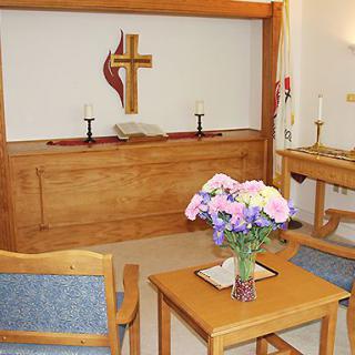 A simple chapel room featuring an altar with a cross, chairs, and a table with flowers.