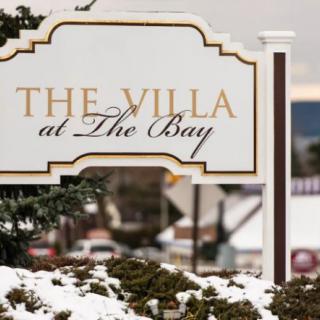 A sign reading 'The Villa at The Bay' surrounded by snow-covered ground and shrubs.