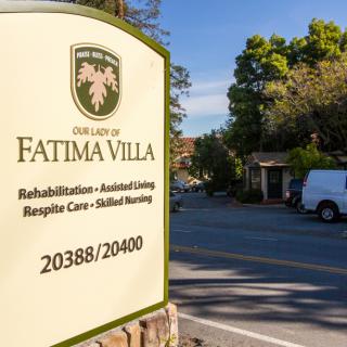 Entrance sign for Fatima Villa on a sunny street with trees and vehicles.