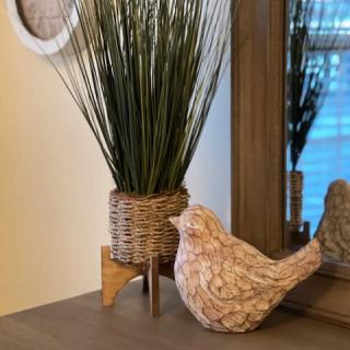 A decorative tabletop display featuring a wicker plant holder with tall grass and a bird sculpture beside a mirror.