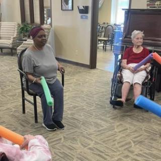 A group of senior citizens participating in a seated exercise session indoors, using pool noodles.