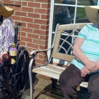 Two elderly individuals sitting outdoors, one in a wheelchair and the other on a bench, both wearing hats.