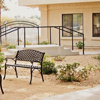 A peaceful outdoor area featuring a metal bench, surrounded by plants and pathways near a building.