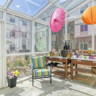 A bright sunroom with a glass ceiling, featuring colorful hanging decorations, a striped chair, and a variety of potted plants.