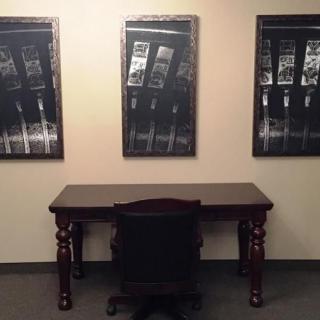A minimalist office setup with a wooden desk, chair, and three framed artworks on the wall.
