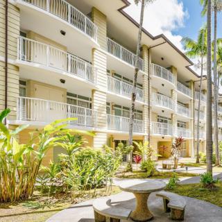 A modern apartment building with balconies, surrounded by palm trees and outdoor seating on a sunny day.