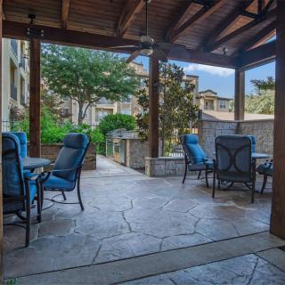 A cozy outdoor patio with seating under a covered pavilion, surrounded by greenery and buildings.