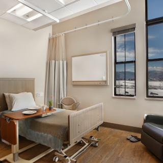 A modern hospital patient room featuring a bed, ample natural light from large windows, and a comfortable sitting area.