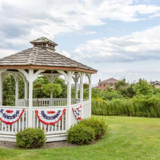A white gazebo decorated with red, white, and blue bunting sits on a lush green lawn under a partly cloudy sky.