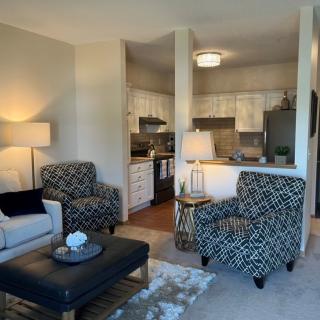 A modern apartment living room with an open kitchen layout, featuring patterned armchairs and ambient lighting.