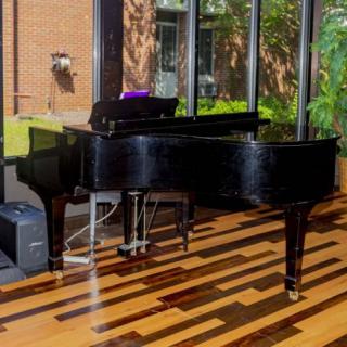 A grand piano positioned in a sunlit room with large windows and a polished hardwood floor.