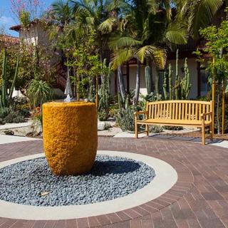 A serene garden setting featuring a central fountain, surrounded by palm trees and complemented by a wooden bench.