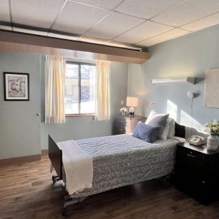 A hospital room with a single bed, neatly arranged with pillows and a blanket, next to a window with curtains. A nightstand holding a lamp and flowers is placed beside the bed.