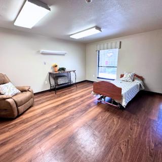A spacious room featuring a bed, a comfortable chair, and wooden flooring with natural light coming through the window.