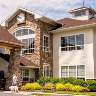 A person walking a dog outside a large residential building with stone and siding exterior.
