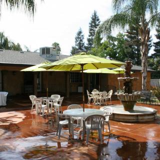 A sunny outdoor patio with tables, chairs, umbrellas, and a central fountain surrounded by palm trees.