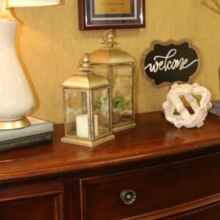 A warmly decorated table featuring a lamp, lanterns with candles, a 'welcome' sign, and a decorative ornament.