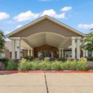A modern building entrance with landscaped surroundings and a driveway under a clear sky.