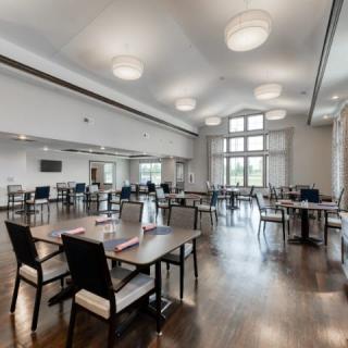 A spacious dining room with wooden floors, well-arranged tables and chairs, featuring large windows and modern lighting.