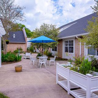 A peaceful courtyard with tables, chairs, and a blue umbrella surrounded by greenery.