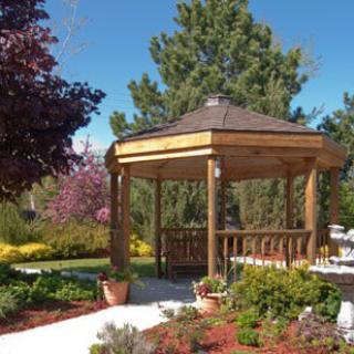 A wooden gazebo surrounded by a landscaped garden with trees and plants, set on a sunny day.