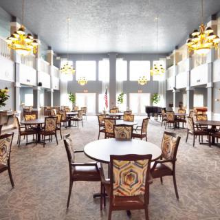 A spacious dining hall with elegant chandeliers and large windows, featuring multiple round tables and chairs arranged neatly.