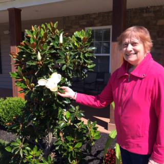 A woman in a pink jacket stands in a garden, smiling and holding a large flower.