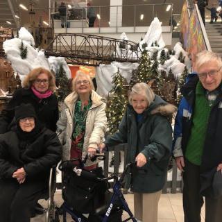 A group of elderly people gathered indoors, dressed in winter clothing and surrounded by holiday decorations.