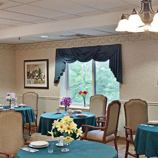 A cozy dining room with tables and chairs set up for a meal, featuring floral centerpieces and a window view.