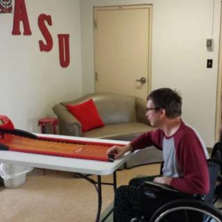 A person in a wheelchair plays shuffleboard indoors near ASU-themed decor.