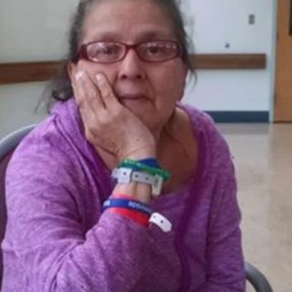 A woman wearing glasses and bracelets sits indoors with her chin resting on her hand.