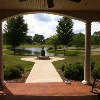 A serene view from a porch overlooking a garden with a statue, pathway, and pond surrounded by trees.