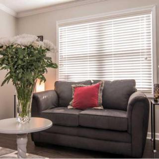 A cozy living room with a dark sofa, decorated with colorful cushions. A vase of flowers sits on the coffee table next to it, and natural light filters through window blinds.