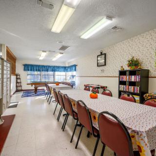 A community center meeting area with a long table covered in a patterned cloth and surrounded by red chairs, featuring a bookshelf and a pool table in the background.