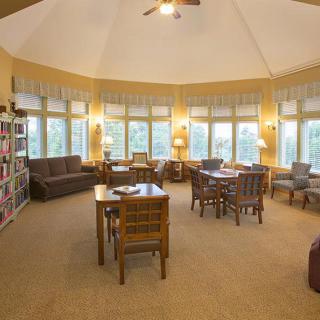 A cozy library reading room with bookshelves, tables, and chairs by large windows.