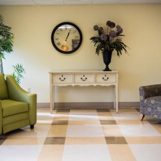 A cozy living room setup featuring a green armchair, a patterned sofa, a decorative table with flowers, and a wall clock.