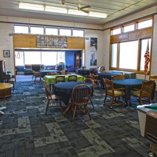 A community hall with several tables and chairs, featuring large windows and a carpeted floor.