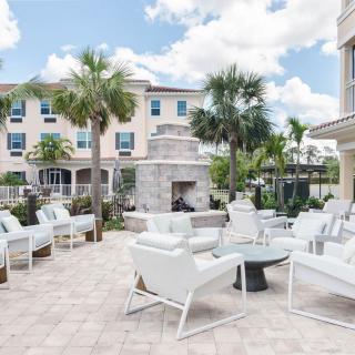A modern outdoor patio area with white furniture and a fireplace, surrounded by palm trees and buildings.