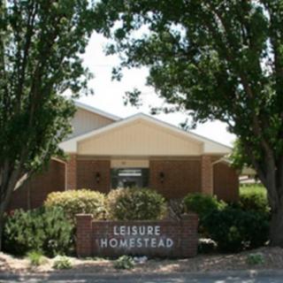 The entrance of a building with a sign reading 'Leisure Homestead' surrounded by trees and shrubs.