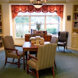 A cozy library reading area with a table, chairs, and stacks of books near a large window.