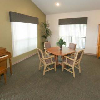A cozy dining area with a round table and four chairs, complemented by large windows with blinds, an upright piano, and a potted plant in the corner.