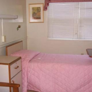 A simple hospital room with a single bed covered in pink bedding, next to a nightstand and a window with curtains.