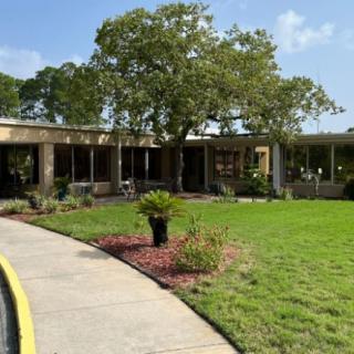 A single-story building surrounded by grass and trees, with a curved pathway leading to the entrance.