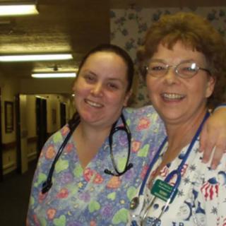 Two smiling nurses in scrubs posing together in a hospital hallway.