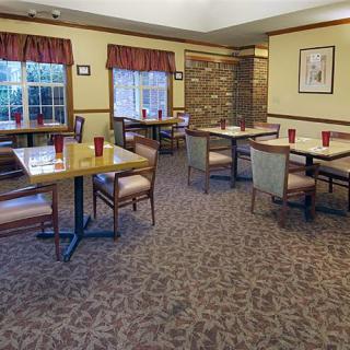 An empty dining room with arranged tables and chairs, featuring carpeted flooring and windows with curtains.
