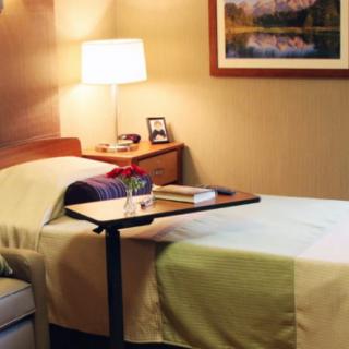 A cozy bedroom or hospital room with a neatly made bed and a bedside table under a warm lamp light.