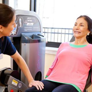 A woman exercises on a leg press machine while being assisted by a personal trainer at the gym.