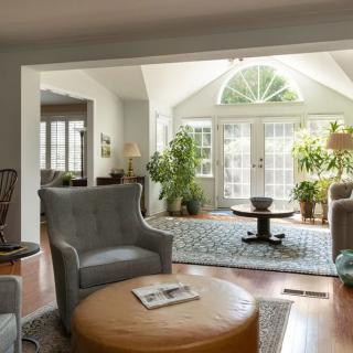 A cozy living room with ample natural light, featuring comfortable armchairs, plants, and a wooden floor.