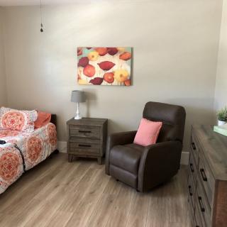 A cozy bedroom with a bed, recliner, and dresser, featuring a floral painting and warm decor.