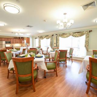 A well-lit dining room with elegantly set tables and chairs, featuring chandeliers and a kitchen area.